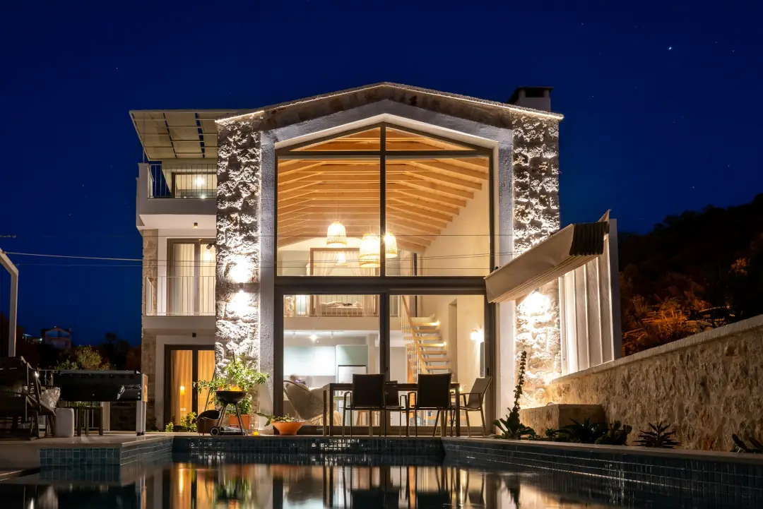 A stone and glass residence lit from within at night, beside its pool